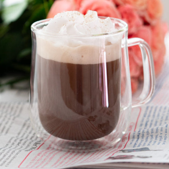 Clear mug with mushroom mocha hot chocolate and whipped cream next to a package of 'Cacao Immortality' coffee on a newspaper.