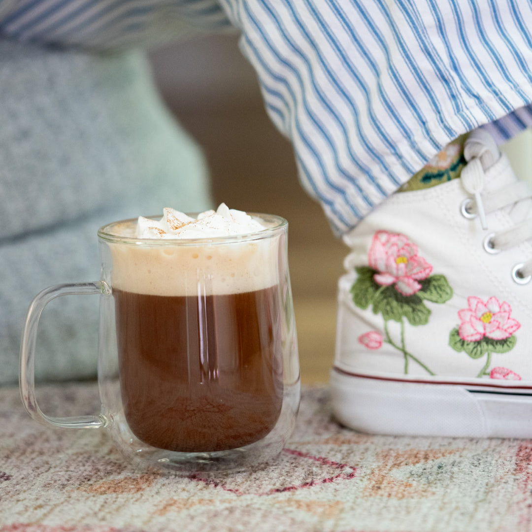 Clear mug with mushroom mocha hot chocolate and whipped cream next to a shoe with floral design on a patterned surface.