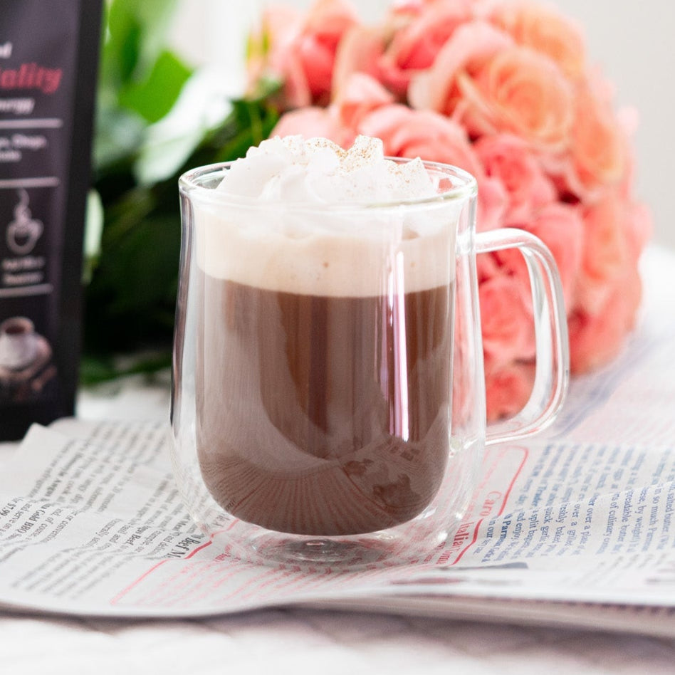 Clear mug with mushroom mocha hot chocolate and whipped cream next to a package of 'Cacao Immortality' coffee on a newspaper.