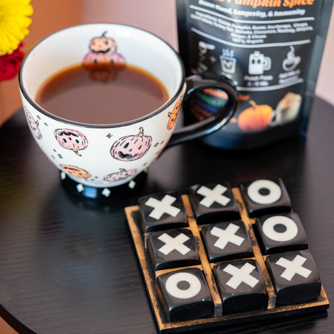 Cup of cacao pumpkin spice, a zplantmagic mushroom koffee drink, with Halloween-themed design, Tic-Tac-Toe game, and pumpkin spice coffee package on a table.